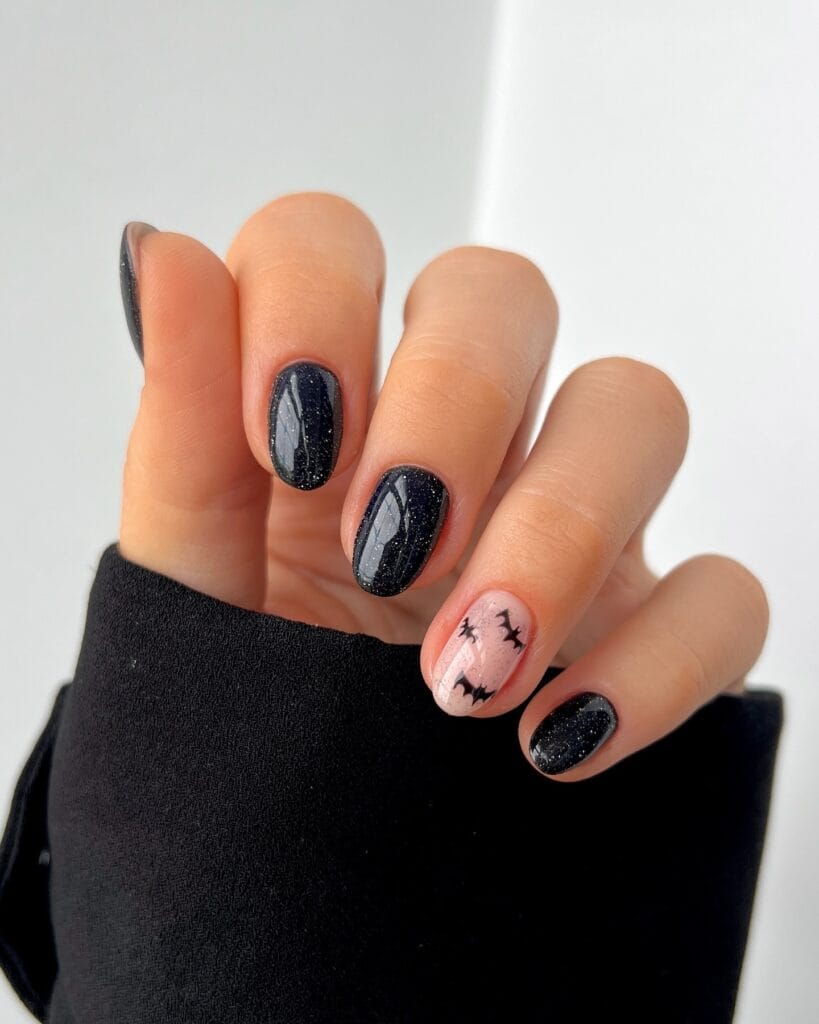 short Halloween nails with black glitter polish and a pale pink accent nail with black bats

