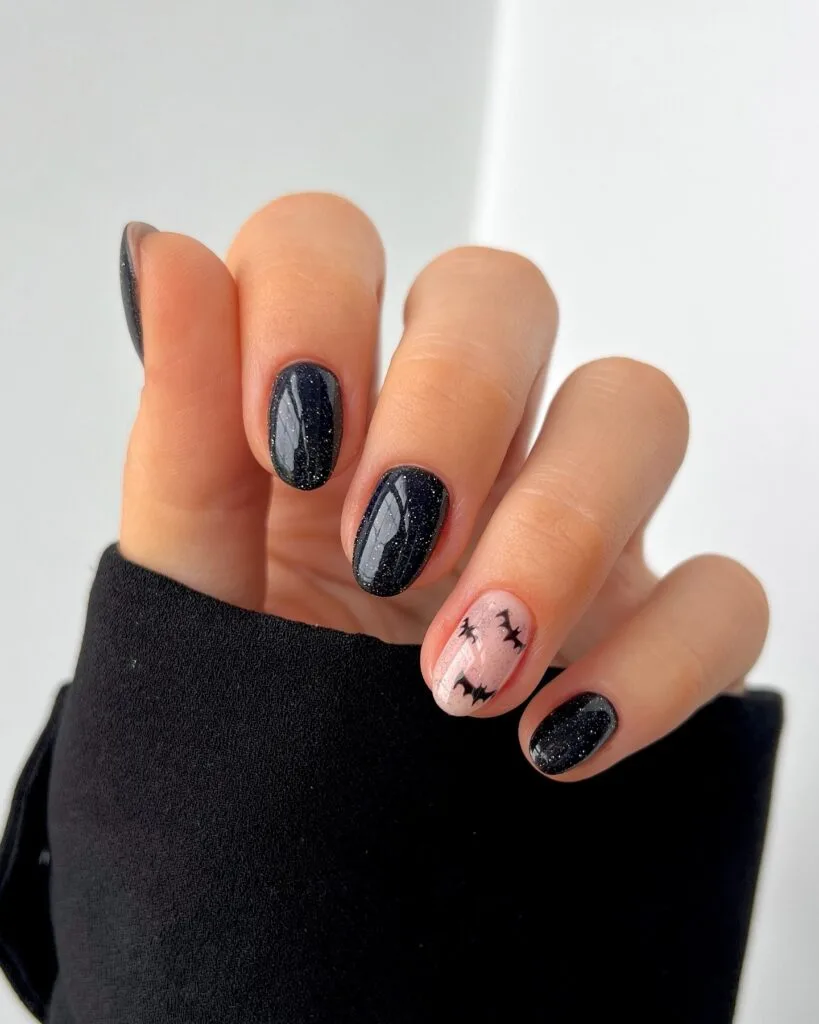 short Halloween nails with black glitter polish and a pale pink accent nail with black bats