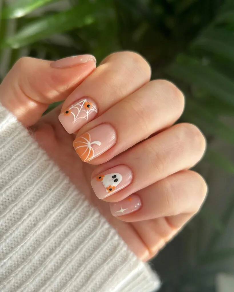 short Halloween nails with nude polish, white spiderweb art, pumpkin detail, and a ghost accent