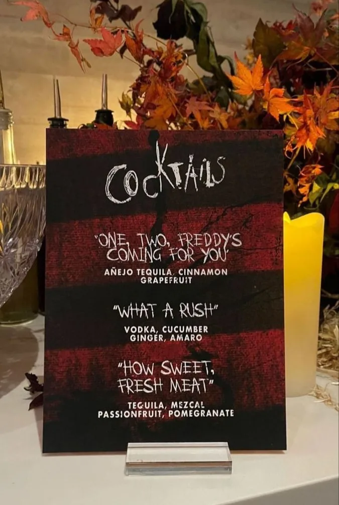 Halloween cocktail menu titled "Cocktails" beside bottles, glassware, candles, and autumn leaves
