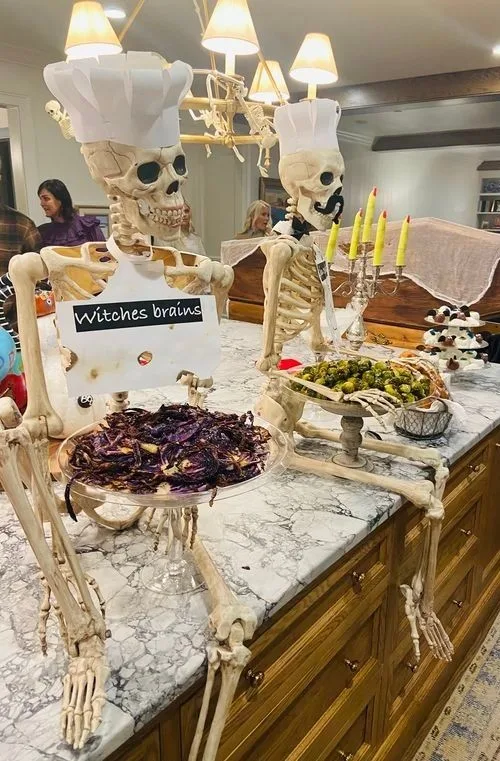Halloween party table with skeleton chefs, candles, food platters, and a "Witches brains" sign

