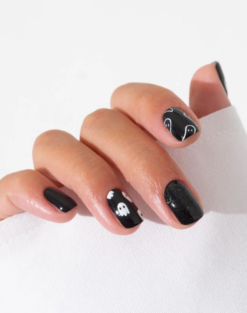 short spooky nails in glossy black with white ghost accents and one black glitter nail