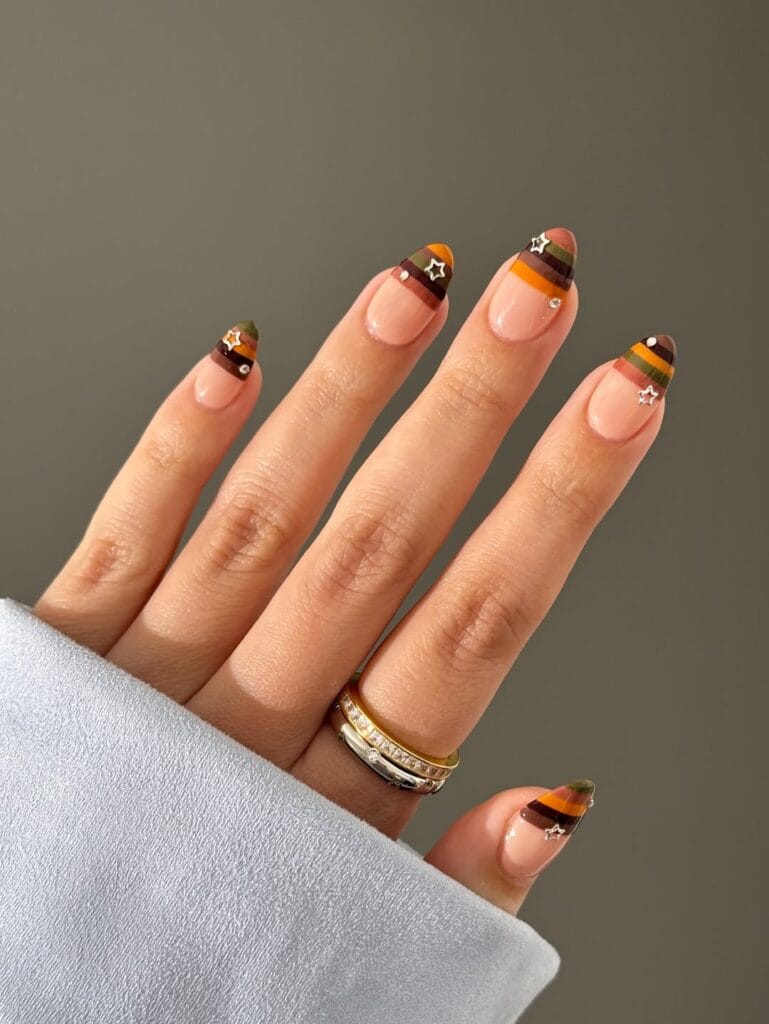 Fall nail designs with striped orange, olive, burgundy, and brown French tips plus silver star accents.
