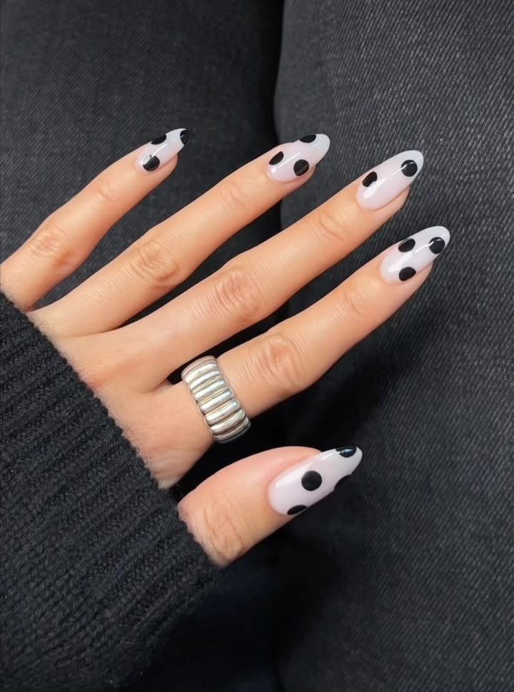 Almond fall polka dot nails with a milky pink-white base and oversized black dots, some cut off at the edges.
