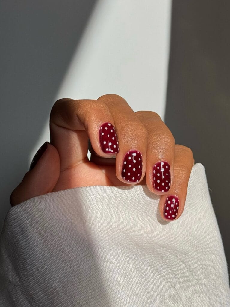 Short, rounded autumn nails with glossy burgundy polish and evenly spaced white polka dots.
