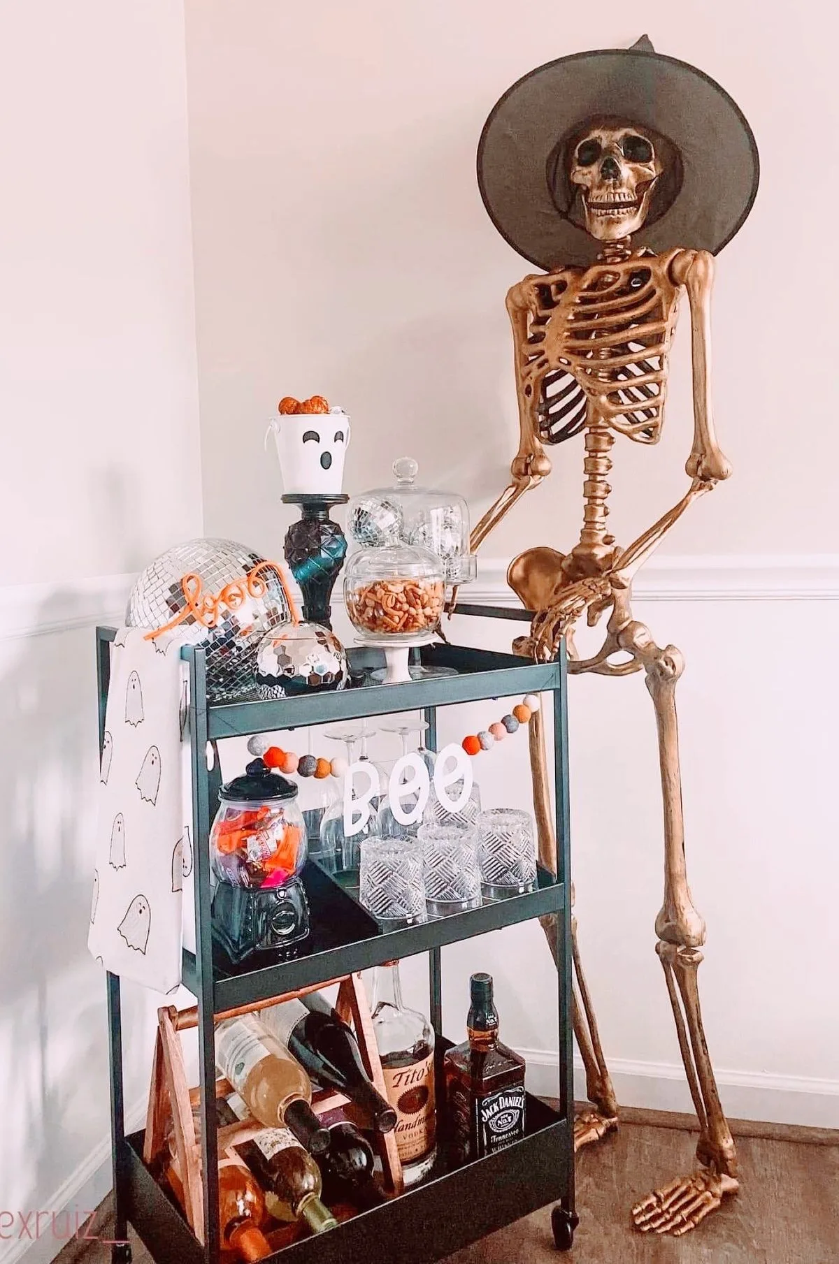 Halloween bar cart with a large skeleton, candy jars, ghost decor, glassware, and liquor bottles
