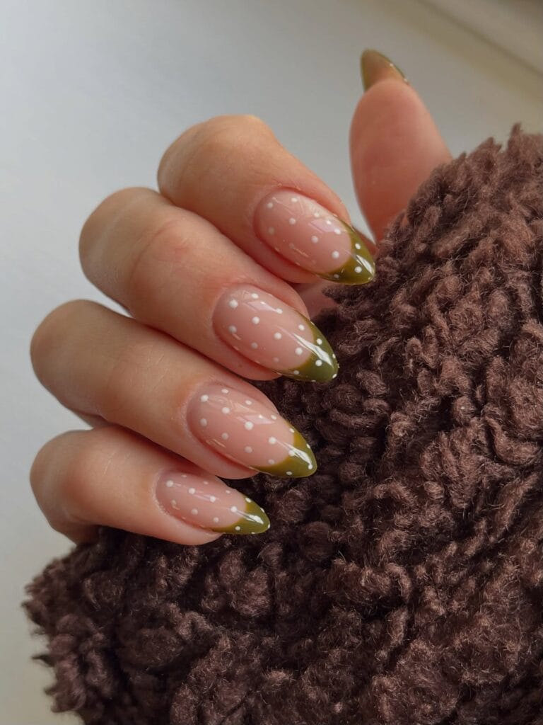 Almond fall polka dot nails with sheer pink bases, olive green French tips, and white dots across both.
