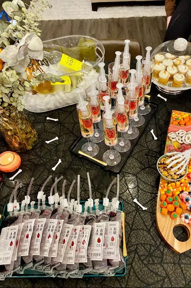 Halloween drink station with syringe shots, faux blood bags marked "A B O," candy, and cupcakes
