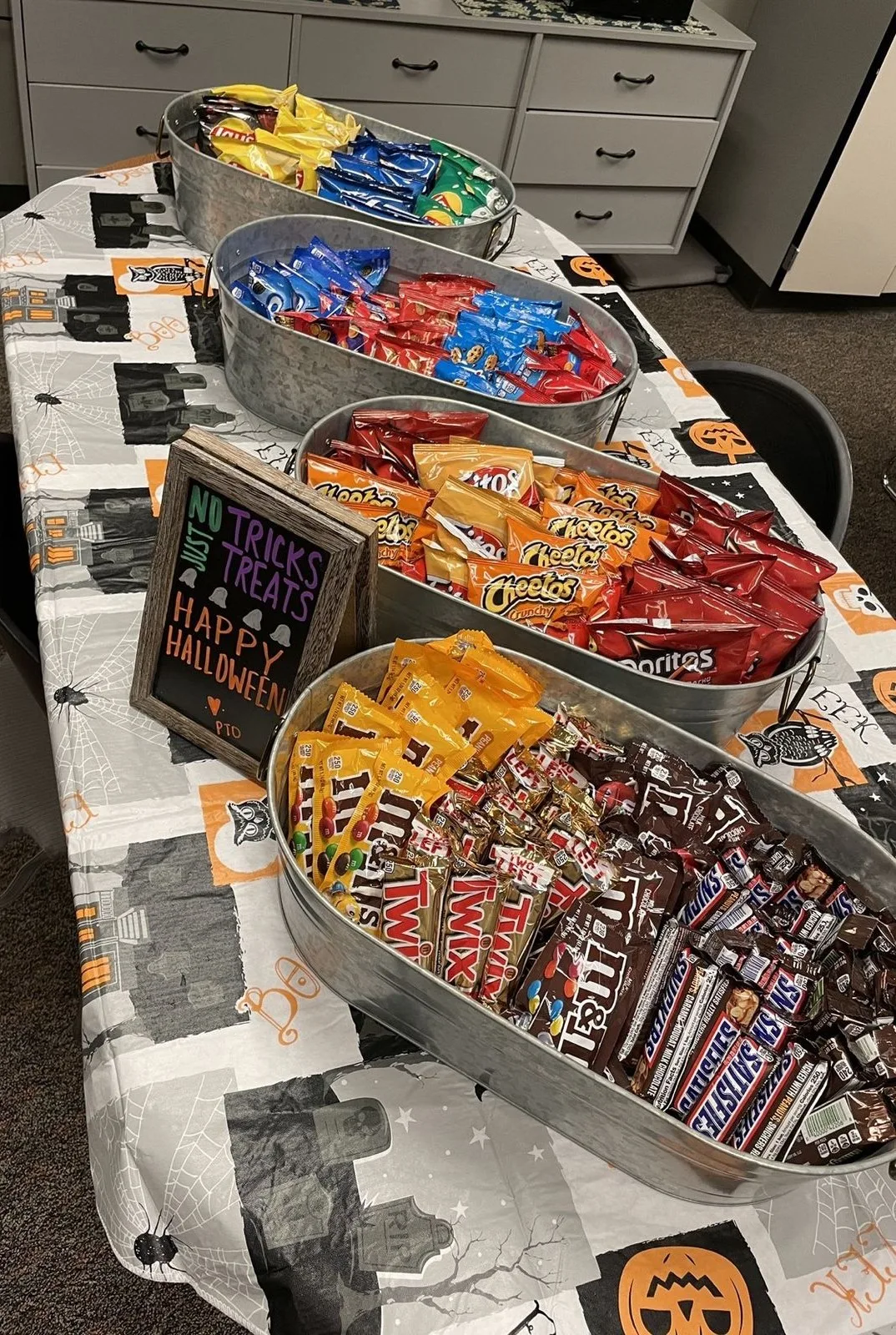 Halloween candy bar with metal tubs of chips and candy beneath a "No tricks just treats" sign
