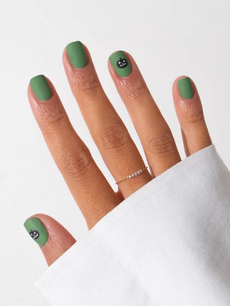 short Halloween nails in matte sage green with small black jack-o’-lantern accents